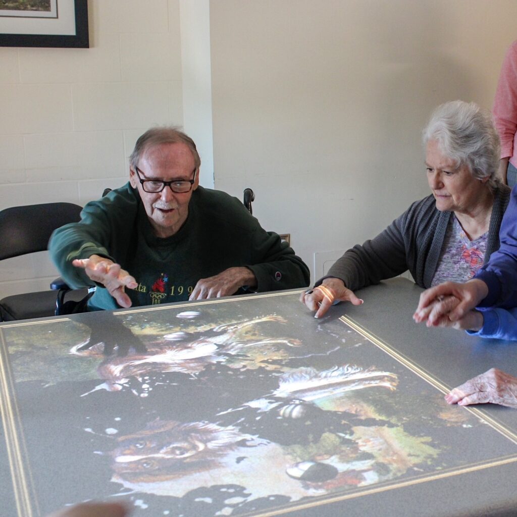 Two older adults gather around a table playing a painting game that is being projected from the Tovertafel system. 